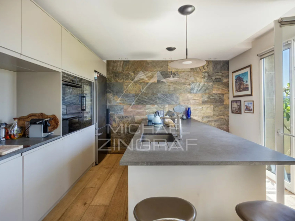 Cuisine moderne avec bar, meubles clairs et mur en pierre, vue sur un espace extérieur.