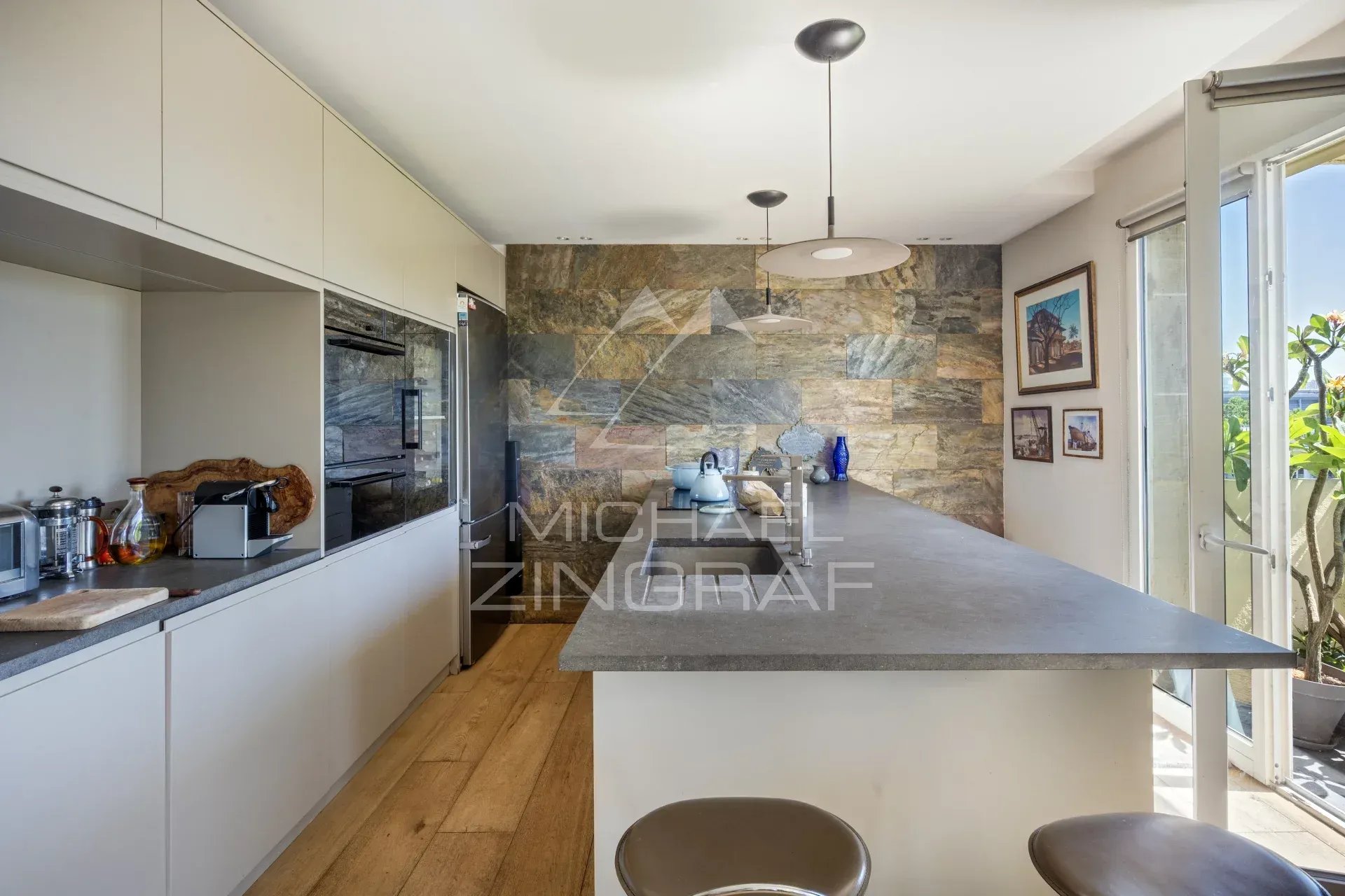 Cuisine moderne avec bar, meubles clairs et mur en pierre, vue sur un espace extérieur.