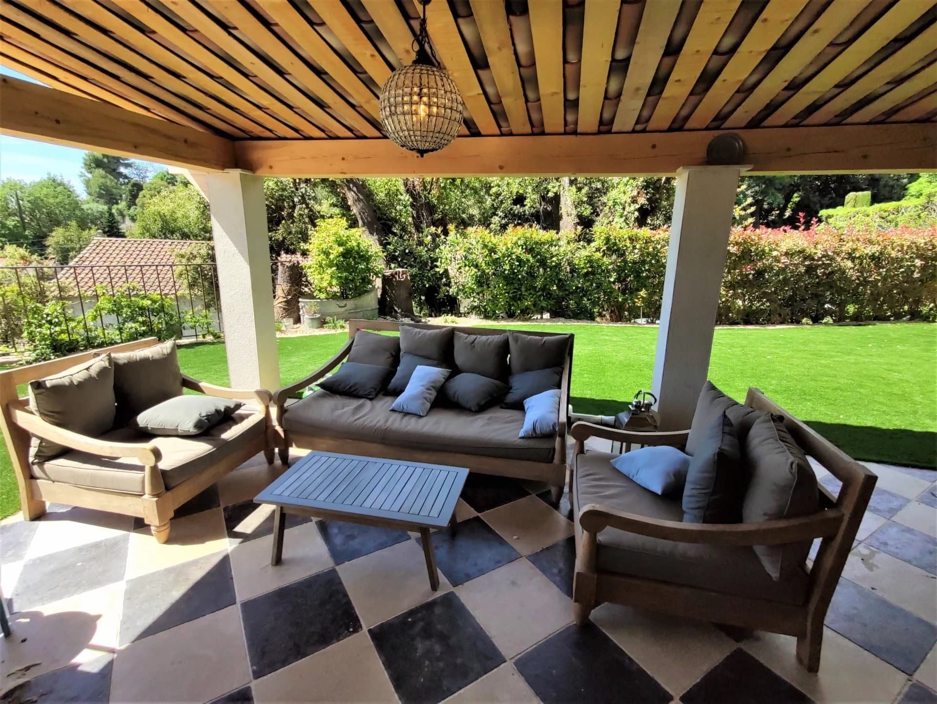 Terrasse avec mobilier en bois et coussins, vue sur jardin verdoyant.