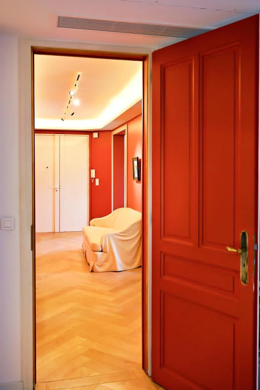 Vue d'un hall d'entrée avec une porte rouge et un salon lumineux en arrière-plan.