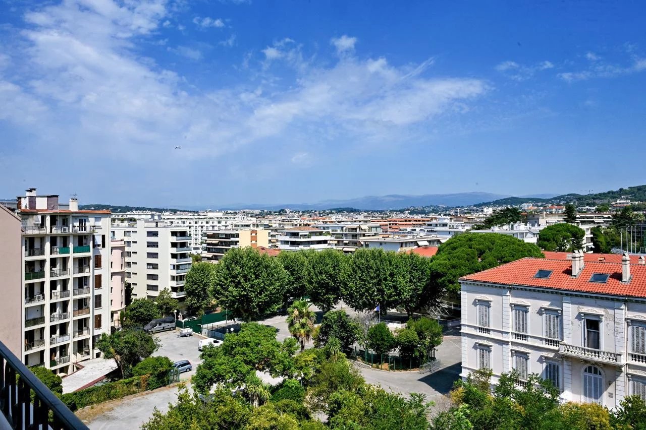 Vue panoramique sur Cannes depuis un appartement bourgeois, avec des bâtiments et des arbres en premier plan.