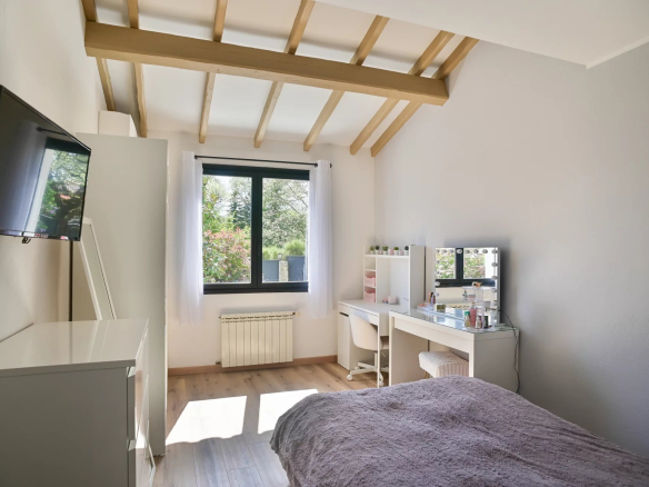 Bright minimalist bedroom with white furniture, a vanity desk and mirror, bed with a gray blanket, and a garden view through a large window.