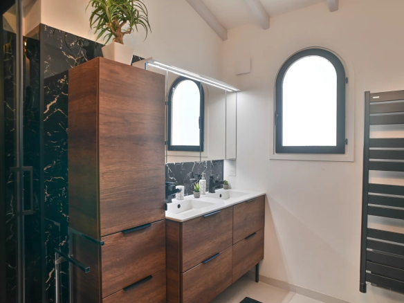 Modern bathroom with a wooden double vanity, white sinks, and a black marble backsplash under mirrored cabinets. A tall plant sits on a cabinet above. Arched window to the right brings in light.