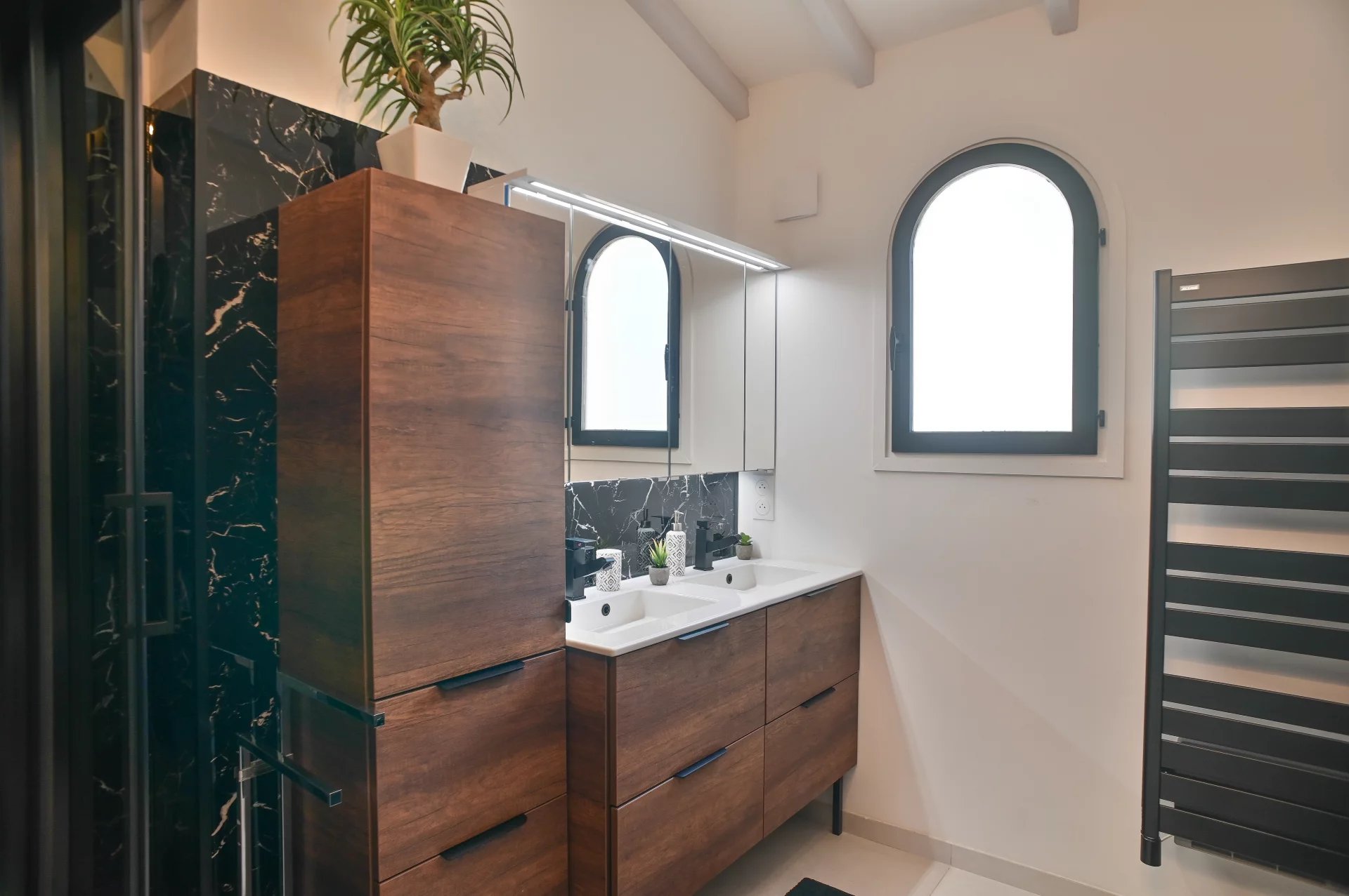 Modern bathroom with a wooden double vanity, white sinks, and a black marble backsplash under mirrored cabinets. A tall plant sits on a cabinet above. Arched window to the right brings in light.