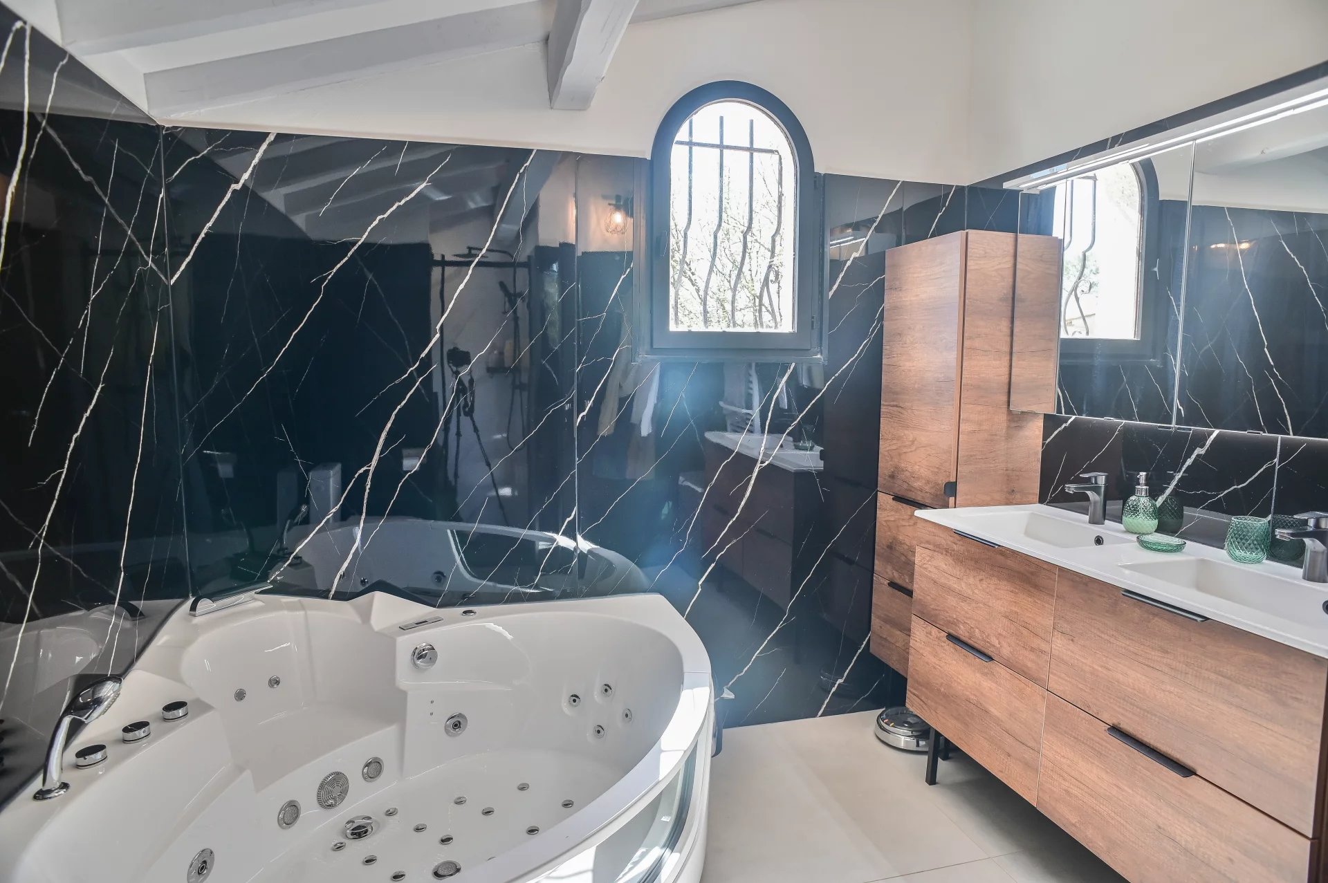 Luxurious bathroom with a white corner whirlpool tub, black veined marble walls, and a wooden double-sink vanity.