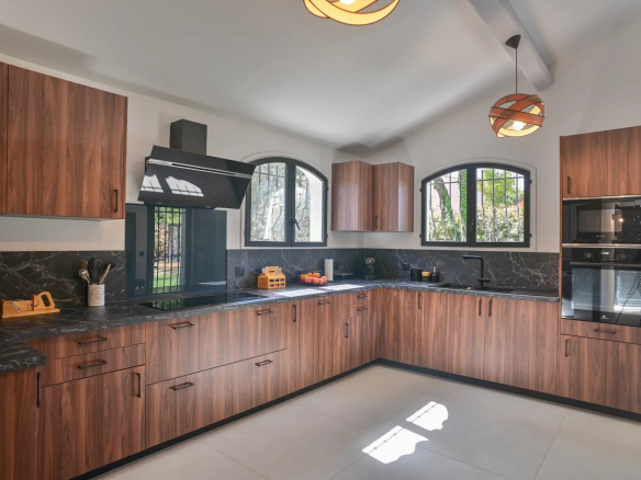 Modern L-shaped kitchen with walnut cabinets, black marble countertops, and a built-in oven along the counter; two arched windows overlook greenery.