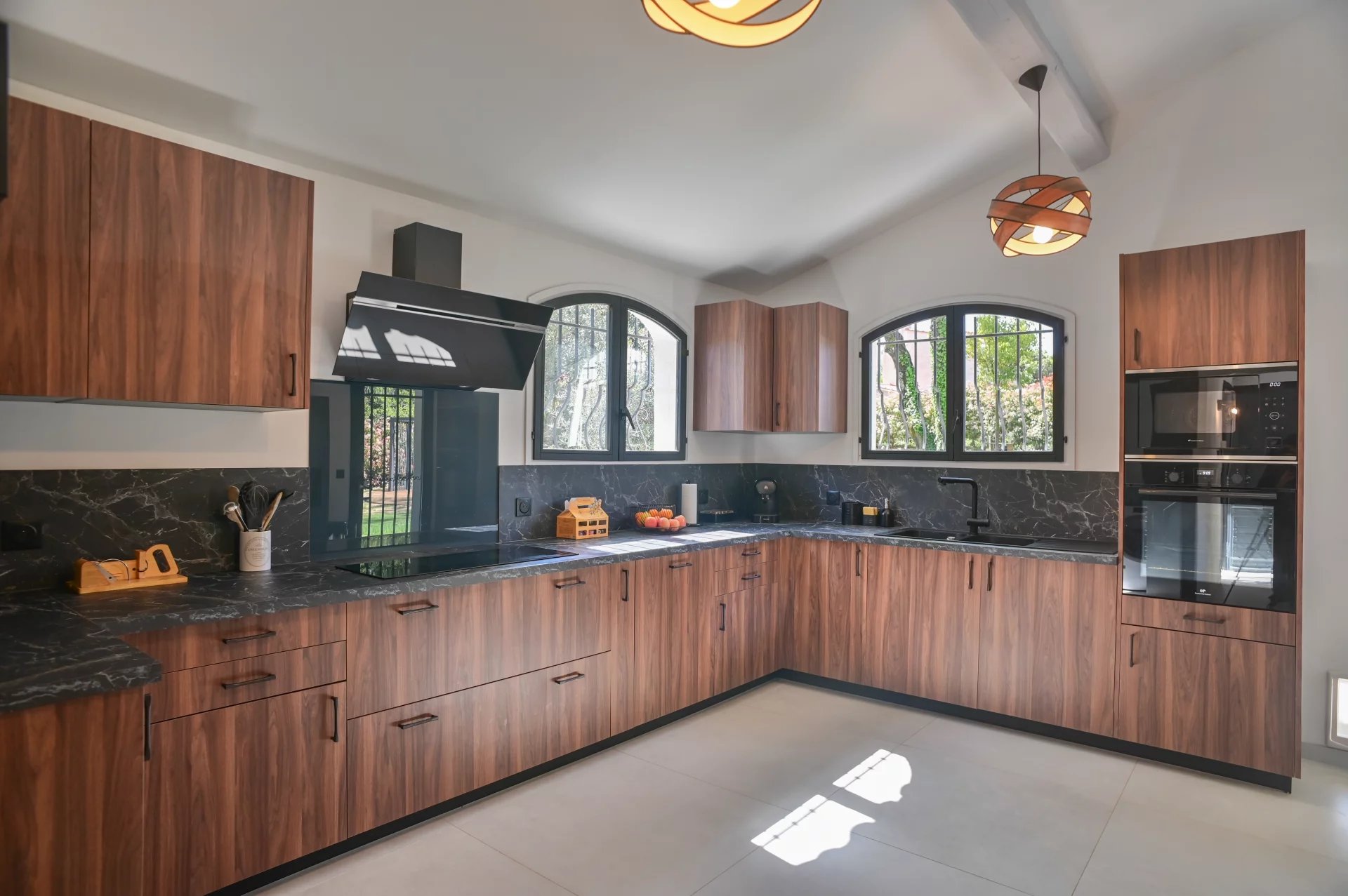 Modern L-shaped kitchen with walnut cabinets, black marble countertops, and a built-in oven along the counter; two arched windows overlook greenery.