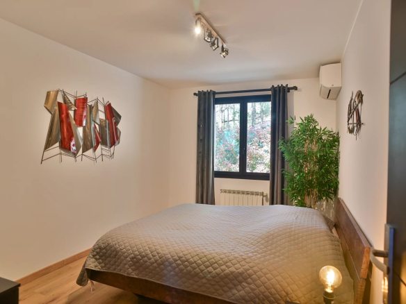 Cozy bedroom with a low wooden bed, grey quilt, and a large window with dark curtains; a tall green plant in the corner.