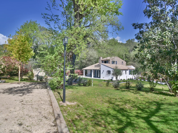 White two-story house with brown-tiled roof in a lush green yard, trees, and a gravel path on a sunny day