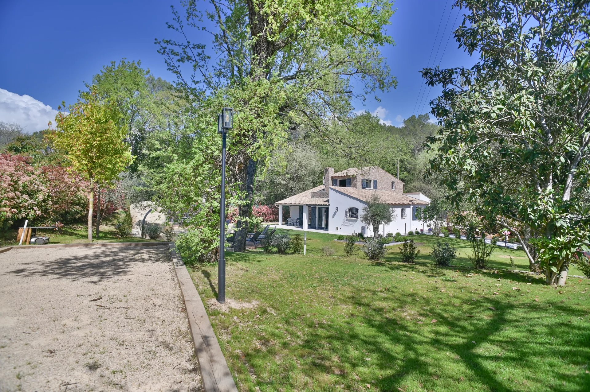 White two-story house with brown-tiled roof in a lush green yard, trees, and a gravel path on a sunny day
