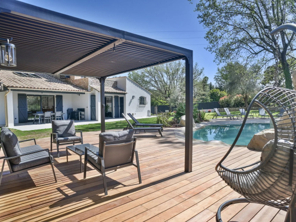 Outdoor backyard with a wooden deck, pool, and gray seating under a pergola near a white house with blue shutters.