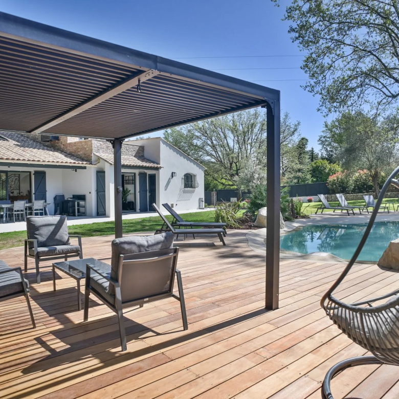 Outdoor backyard with a wooden deck, pool, and gray seating under a pergola near a white house with blue shutters.