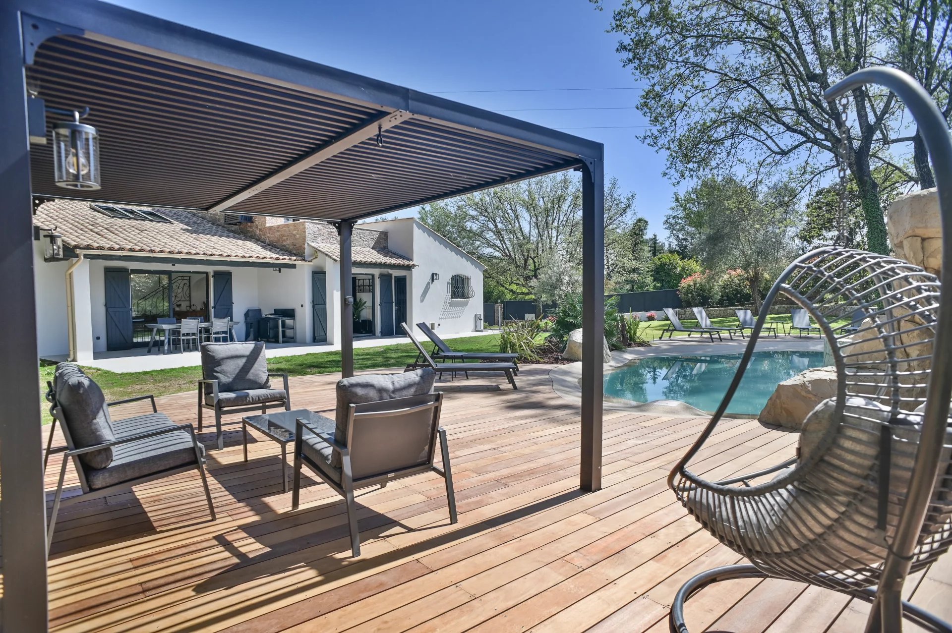 Outdoor backyard with a wooden deck, pool, and gray seating under a pergola near a white house with blue shutters.