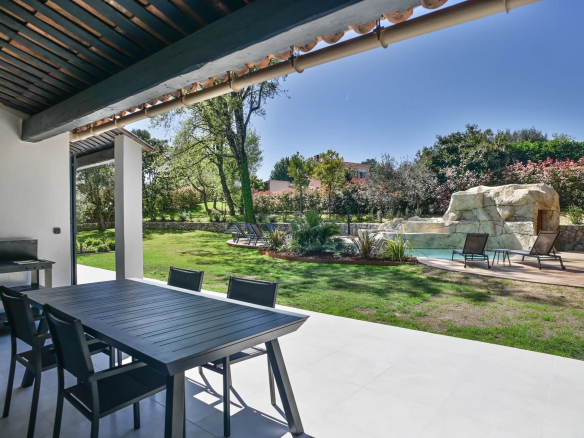 Outdoor patio with a dark dining table and six chairs under a shaded tiled roof, overlooking a green lawn and garden surface.