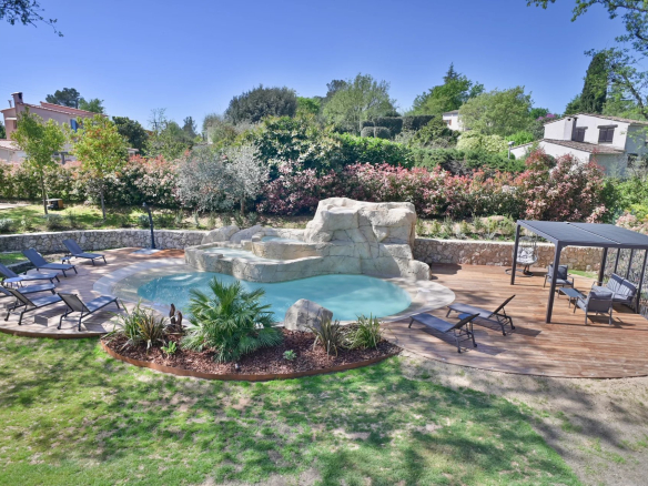 Backyard pool area with rock waterfall, circular wooden deck, and multiple lounge chairs surrounding clear water.