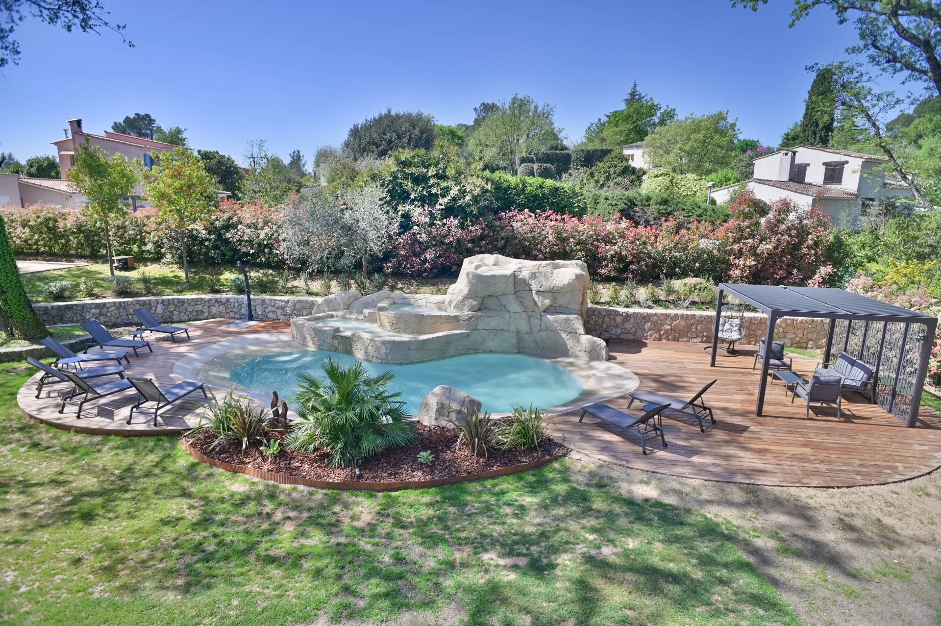 Backyard pool area with rock waterfall, circular wooden deck, and multiple lounge chairs surrounding clear water.