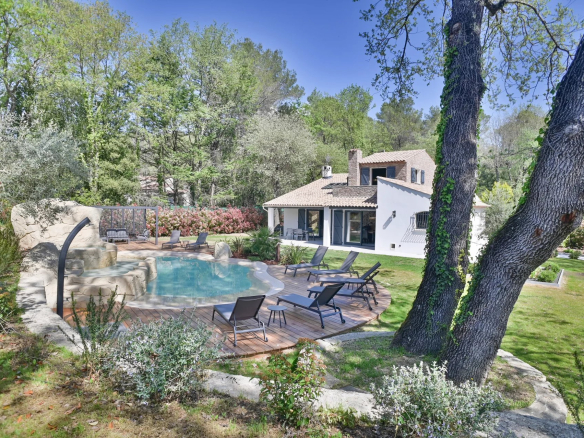 Resort-style backyard with a freeform pool, wooden deck, and lounge chairs beside a white stucco house amid green trees.