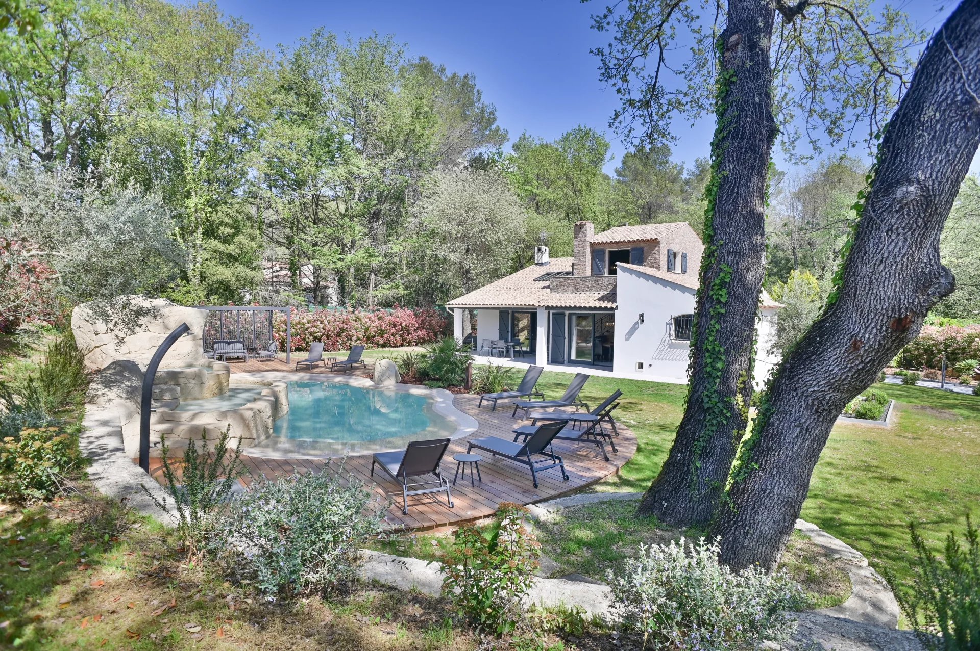 Resort-style backyard with a freeform pool, wooden deck, and lounge chairs beside a white stucco house amid green trees.