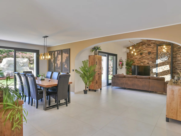 Open-plan dining and living room with a wooden table and black leather chairs, large glass doors to a garden, and an archway leading to a stone-clad TV wall area.