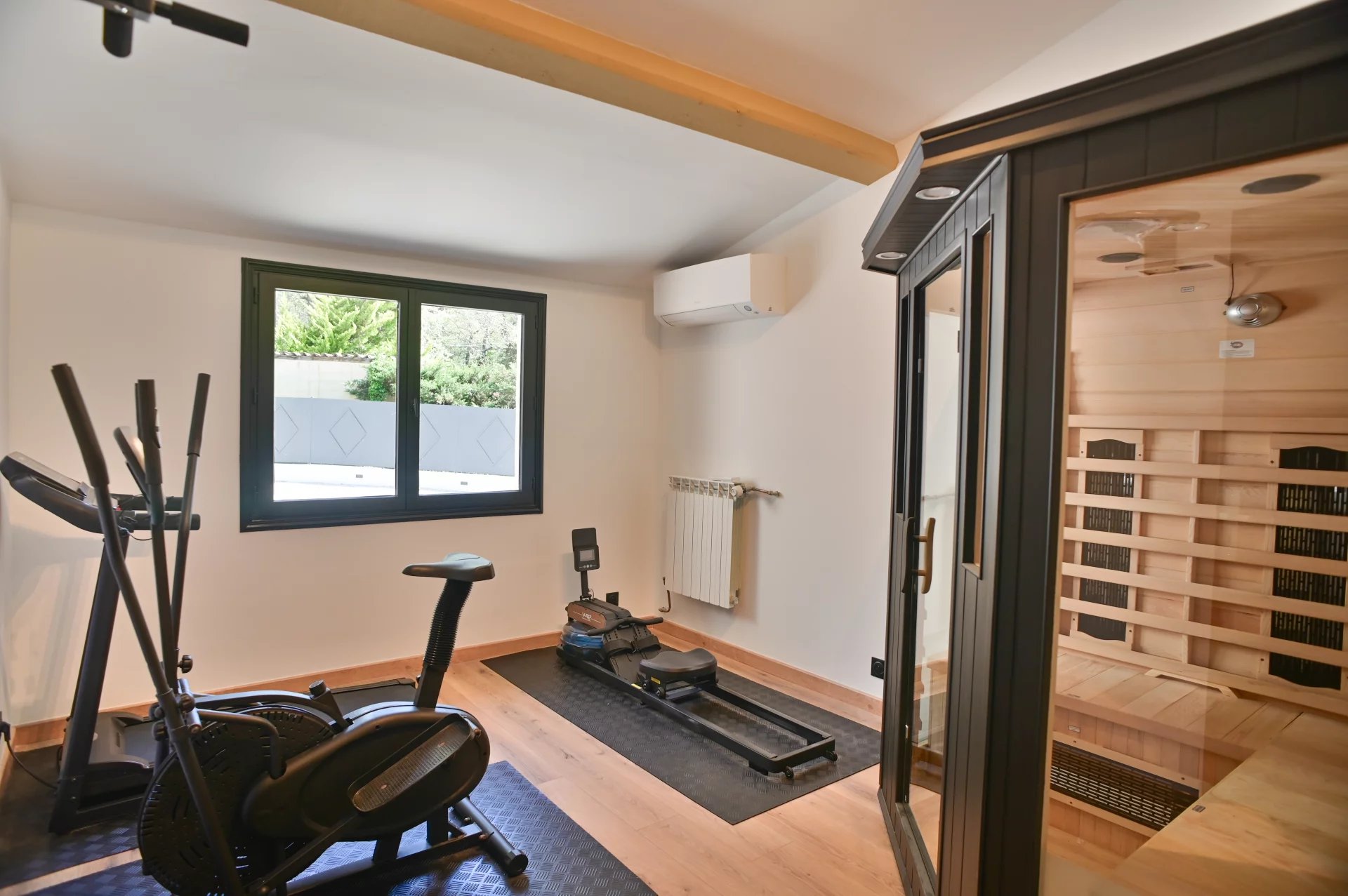 Home gym with an elliptical trainer and a rowing machine on black mats, a window view outside, and a wooden-sauna room behind glass doors to the right.