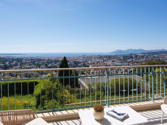 Balcony railing overlooking a coastal city with buildings, blue sea, and distant hills under a clear sky. A white table and chairs sit in the foreground.