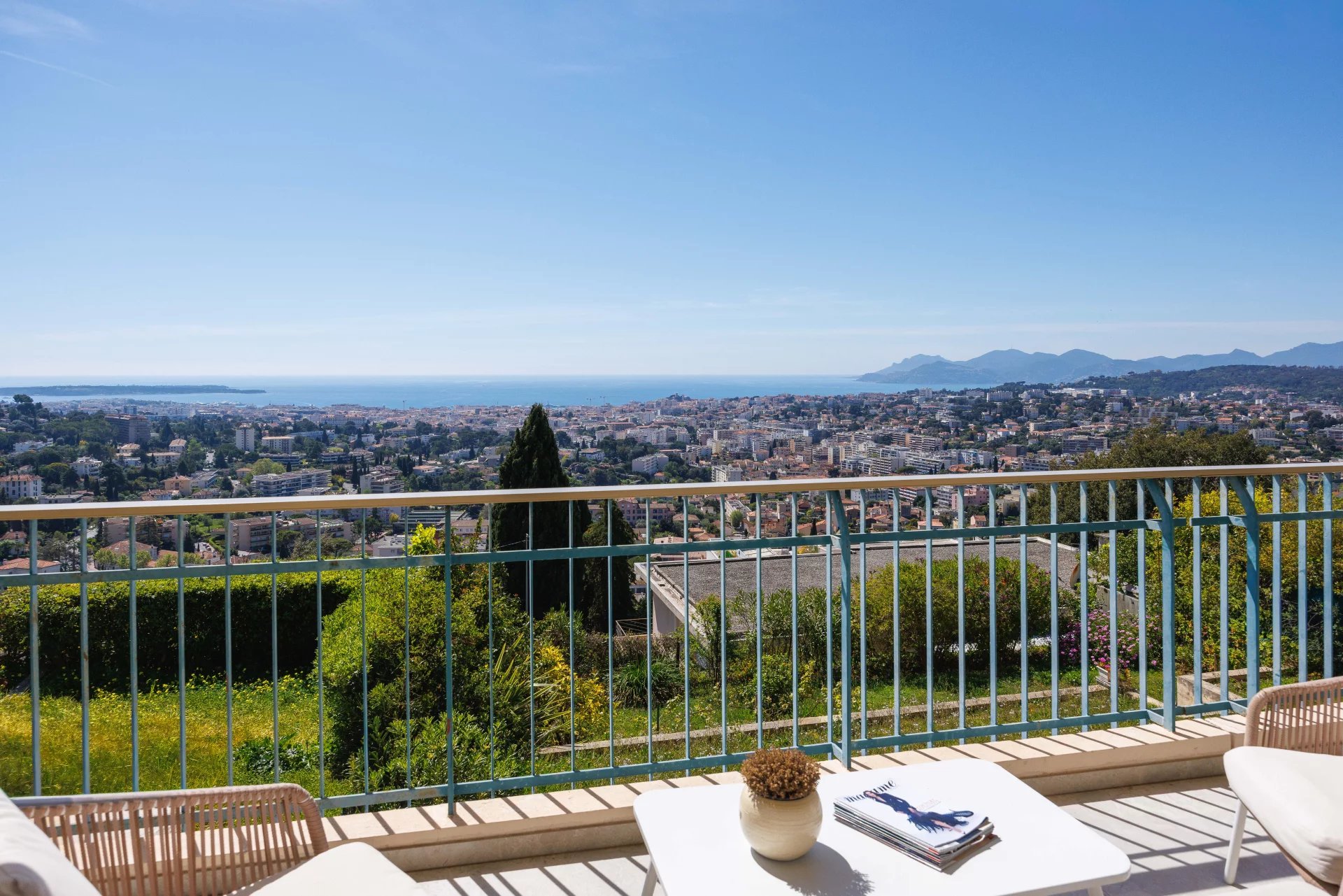 Balcony railing overlooking a coastal city with buildings, blue sea, and distant hills under a clear sky. A white table and chairs sit in the foreground.