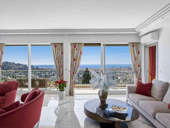 Bright living room with floor-to-ceiling windows showing a coastal city and ocean view; red chairs and beige sofa with red cushions, glass coffee table, and floral curtains.