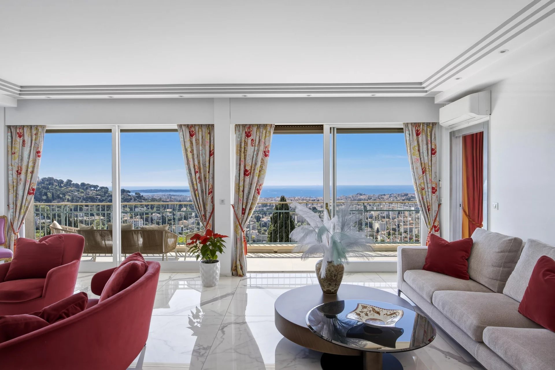 Bright living room with floor-to-ceiling windows showing a coastal city and ocean view; red chairs and beige sofa with red cushions, glass coffee table, and floral curtains.