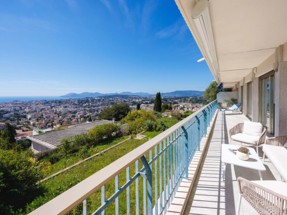 Balcony with white outdoor furniture overlooking a sunny hillside town and distant sea under a blue sky, framed by a blue railing.