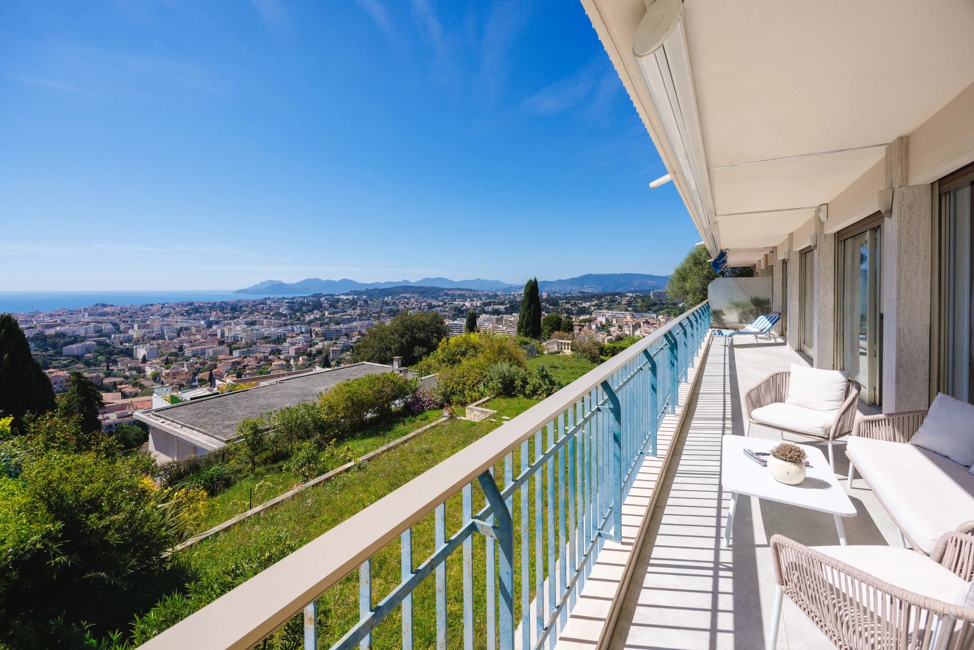 Balcony with white outdoor furniture overlooking a sunny hillside town and distant sea under a blue sky, framed by a blue railing.