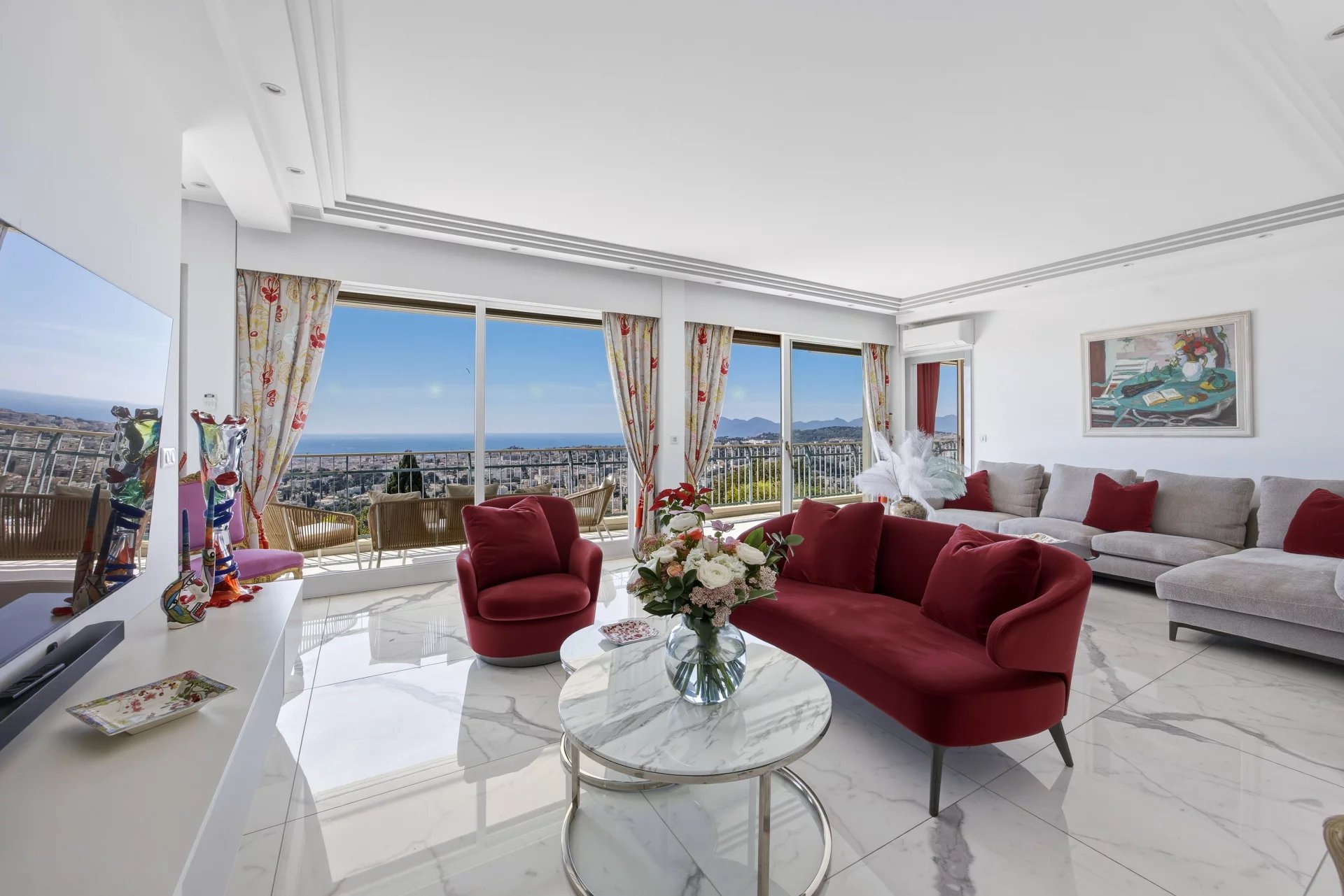 Bright living room with white marble floors, a red velvet sofa and chairs, and a panoramic ocean view through large windows.