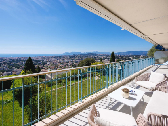 Balcony with white outdoor furniture overlooking a city and coastline under a clear blue sky