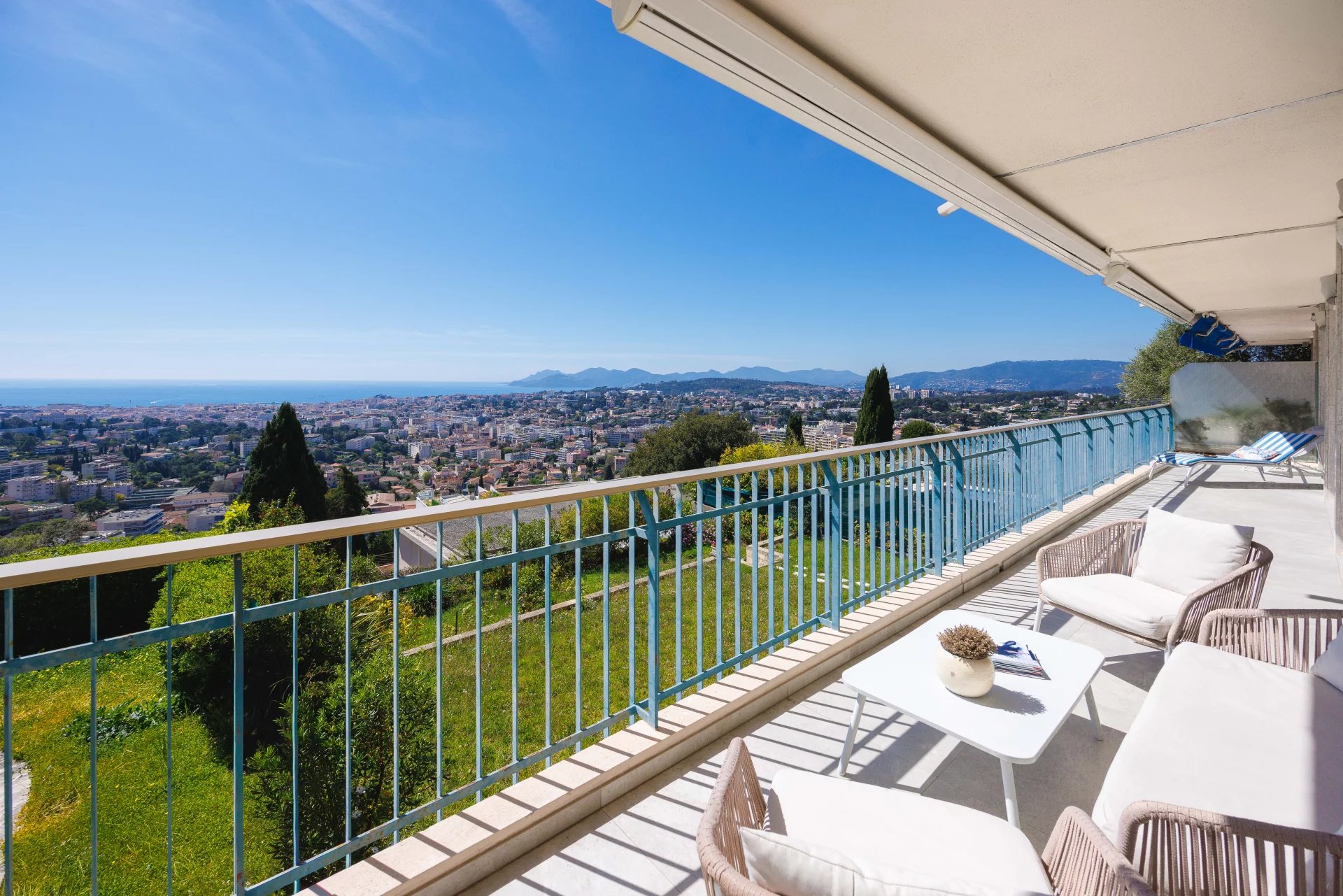 Balcony with white outdoor furniture overlooking a city and coastline under a clear blue sky