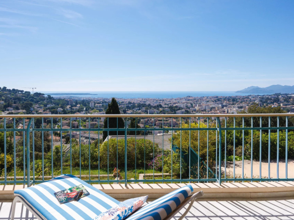 Balcony overlooking a coastal city with a blue railing, a striped blue-and-white lounge chair, and a magazine on the chair, sea in the distance under a clear blue sky.