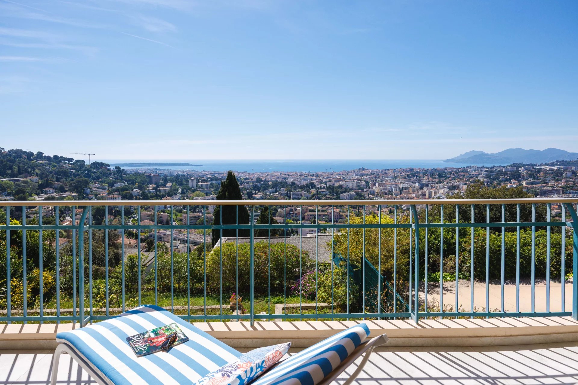 Balcony overlooking a coastal city with a blue railing, a striped blue-and-white lounge chair, and a magazine on the chair, sea in the distance under a clear blue sky.