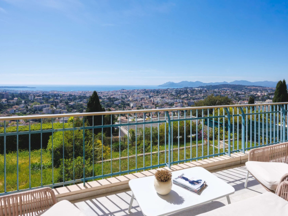 Balcony with a view over a coastal city, sea and distant mountains on the horizon, white table and chairs in the foreground.