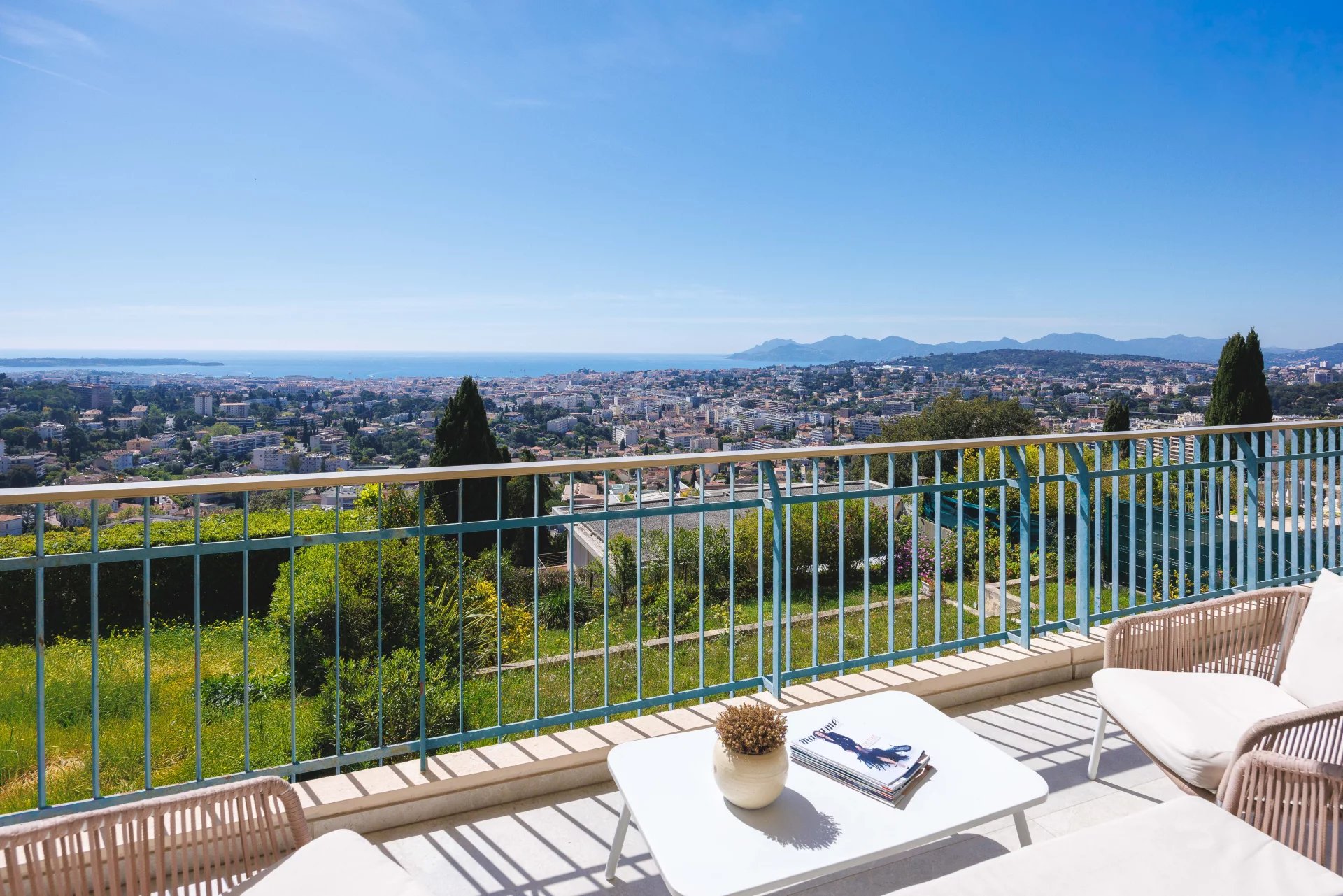 Balcony with a view over a coastal city, sea and distant mountains on the horizon, white table and chairs in the foreground.
