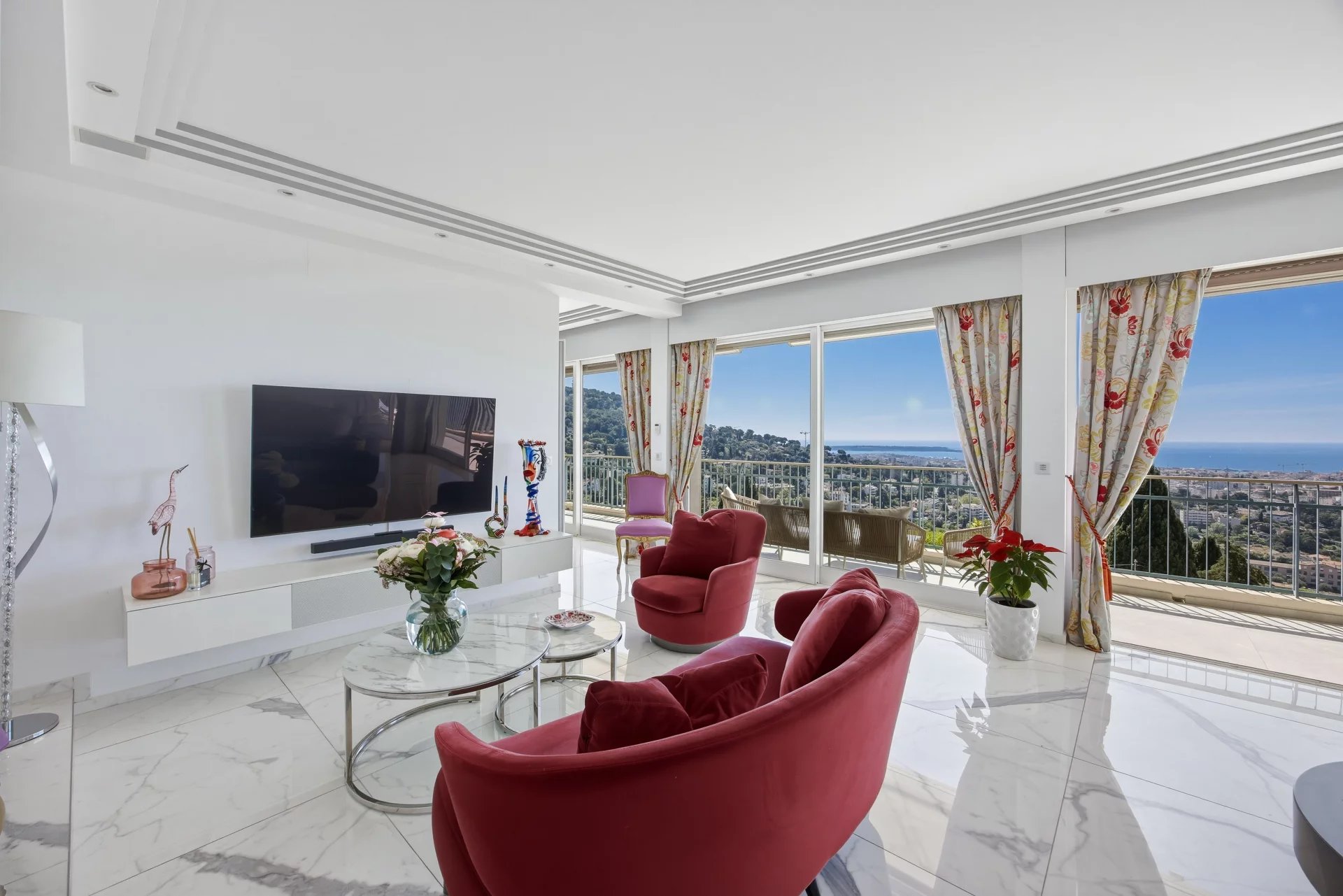 Bright living room with red velvet lounge chairs, marble floor, and a sea-view balcony through large windows.