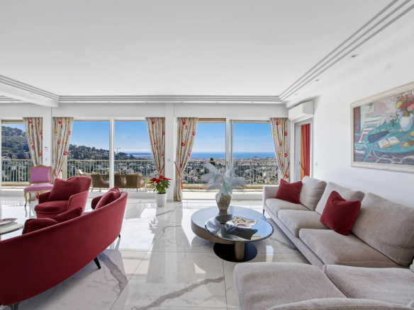 Bright living room with sea view through large windows, beige sectional sofa with red cushions, and red accent chairs around a glass coffee table on a white marble floor with floral curtains and a painting on the wall.