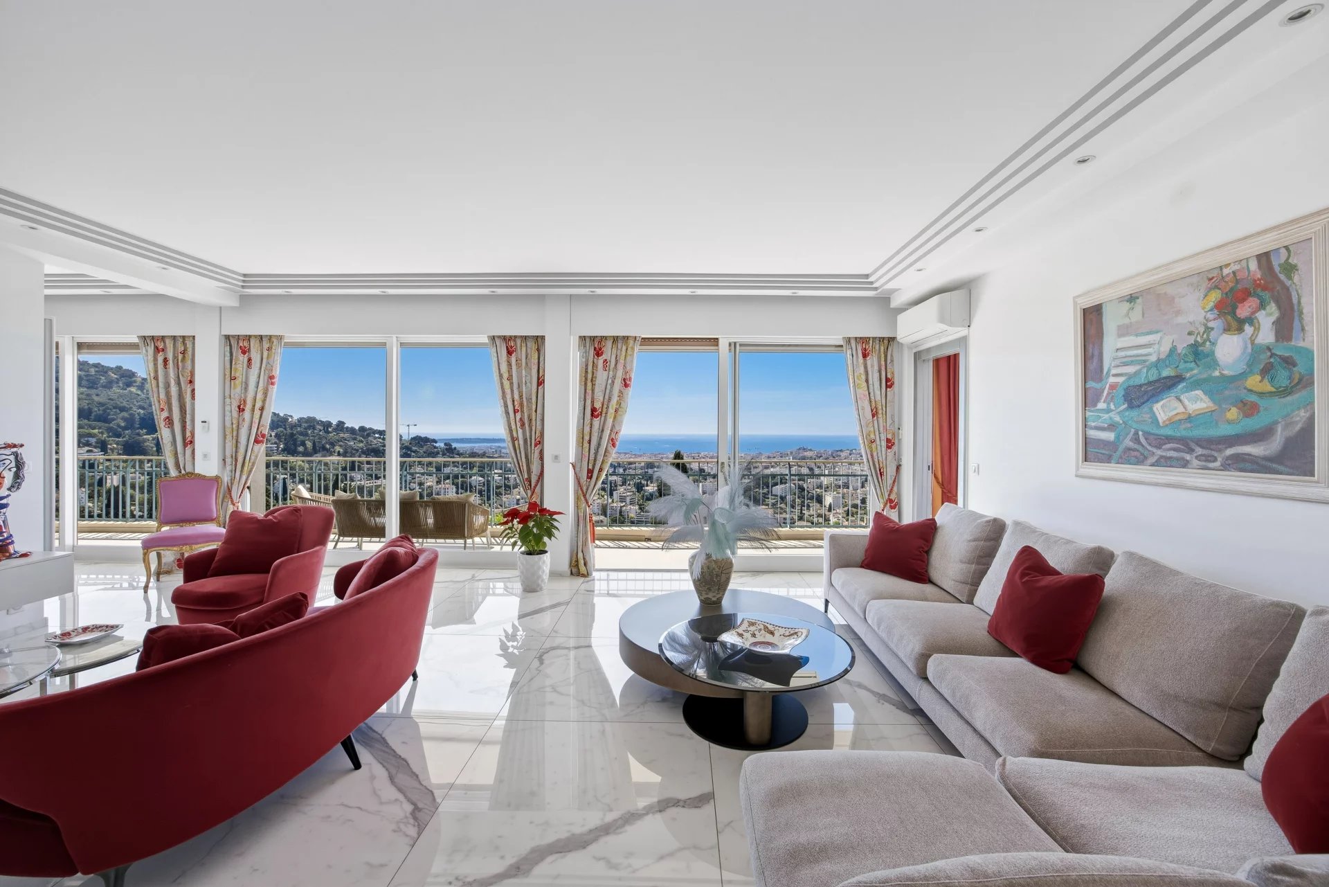 Bright living room with sea view through large windows, beige sectional sofa with red cushions, and red accent chairs around a glass coffee table on a white marble floor with floral curtains and a painting on the wall.