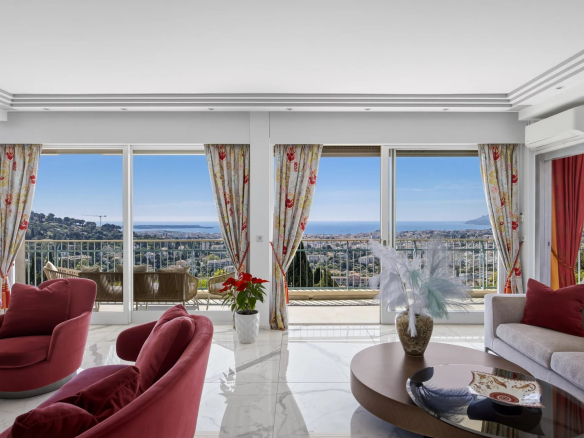Living room with red chairs and cream sofa, overlooking a balcony and an ocean view through large glass doors.