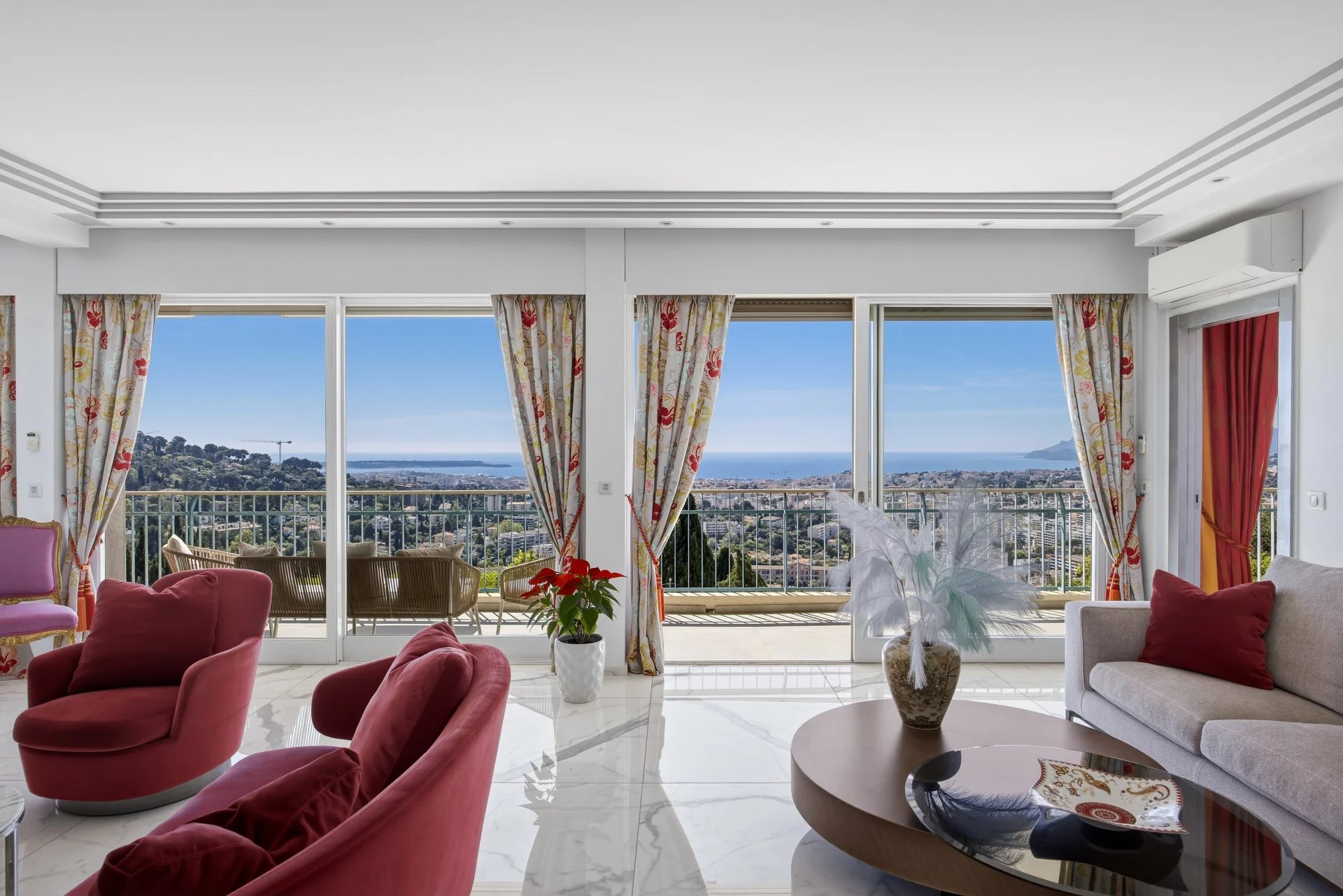 Living room with red chairs and cream sofa, overlooking a balcony and an ocean view through large glass doors.