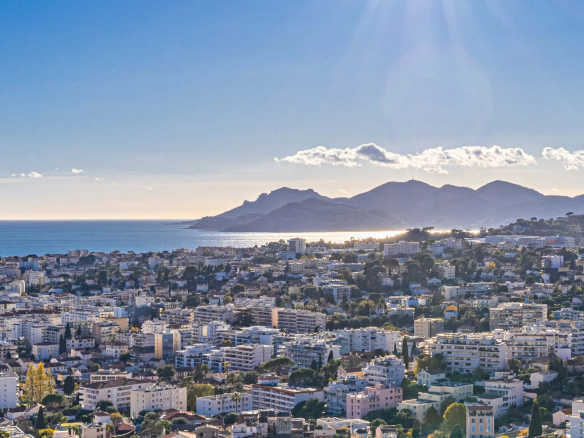 Panoramic view of a seaside city with numerous white apartment buildings, stretching toward a calm blue bay and distant hills under a bright, sunny sky.