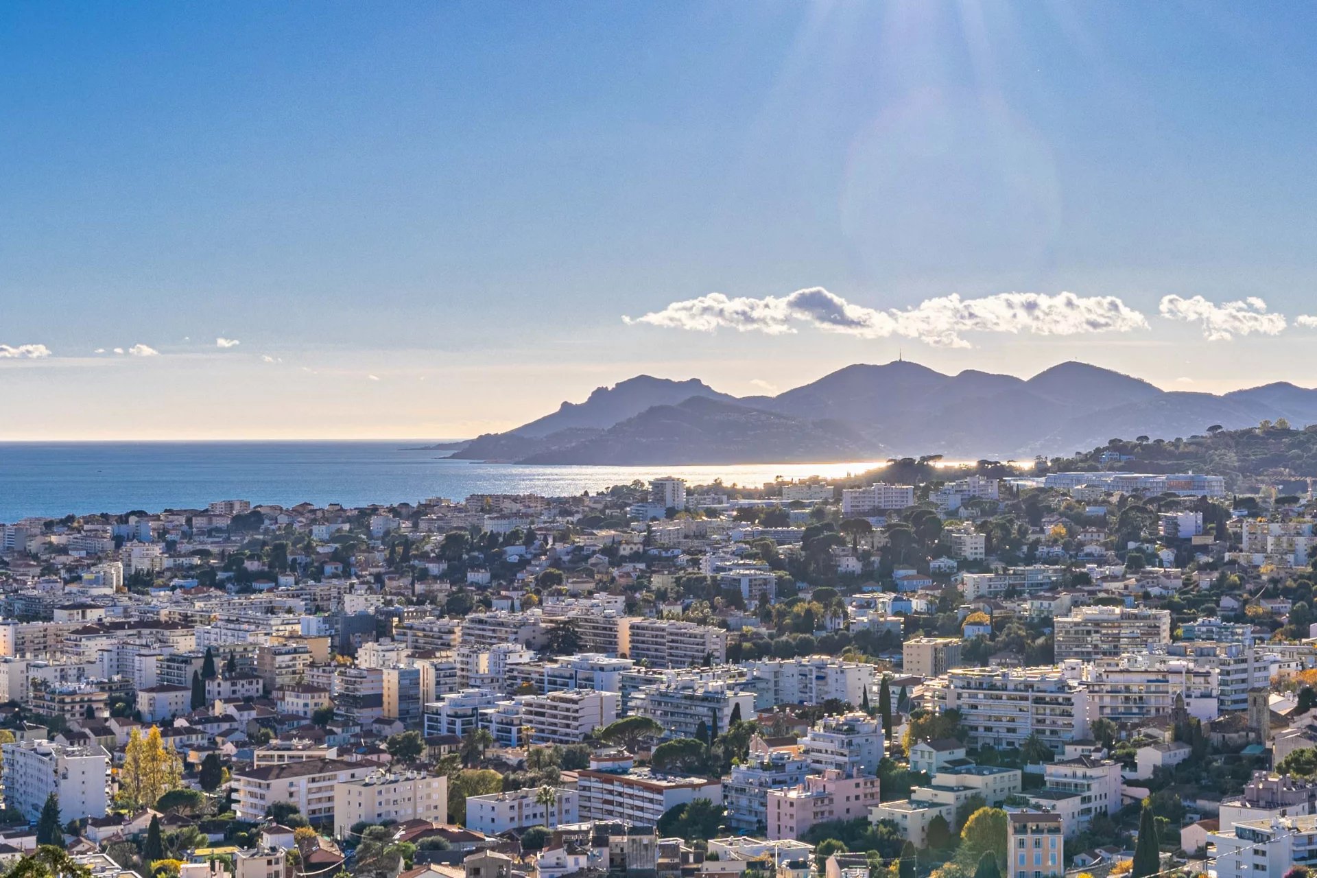 Panoramic view of a seaside city with numerous white apartment buildings, stretching toward a calm blue bay and distant hills under a bright, sunny sky.