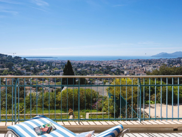 Balcony railing overlooks a coastal city with the sea and mountains in the distance; a striped lounge chair and magazine sit in the foreground.