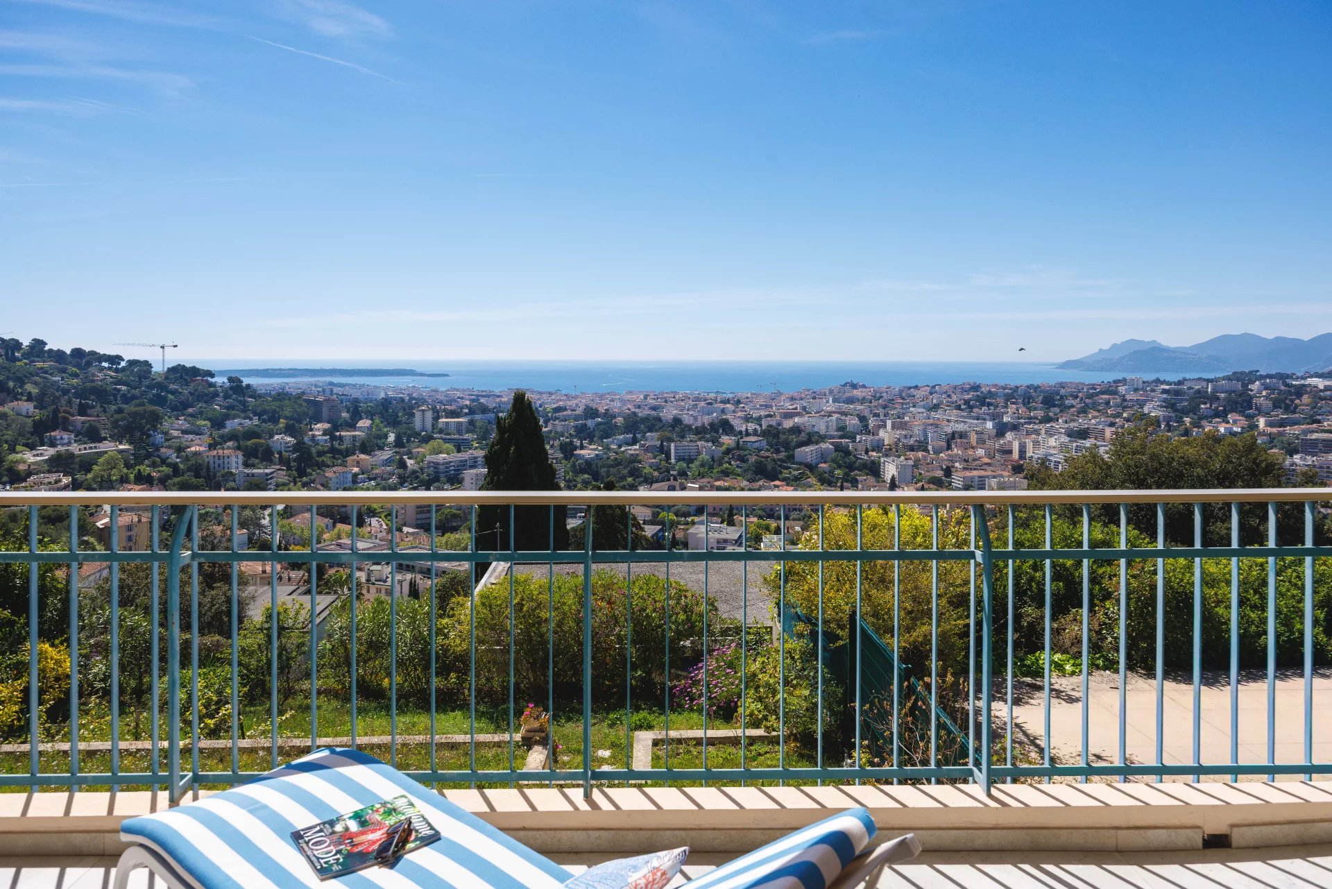 Balcony railing overlooks a coastal city with the sea and mountains in the distance; a striped lounge chair and magazine sit in the foreground.