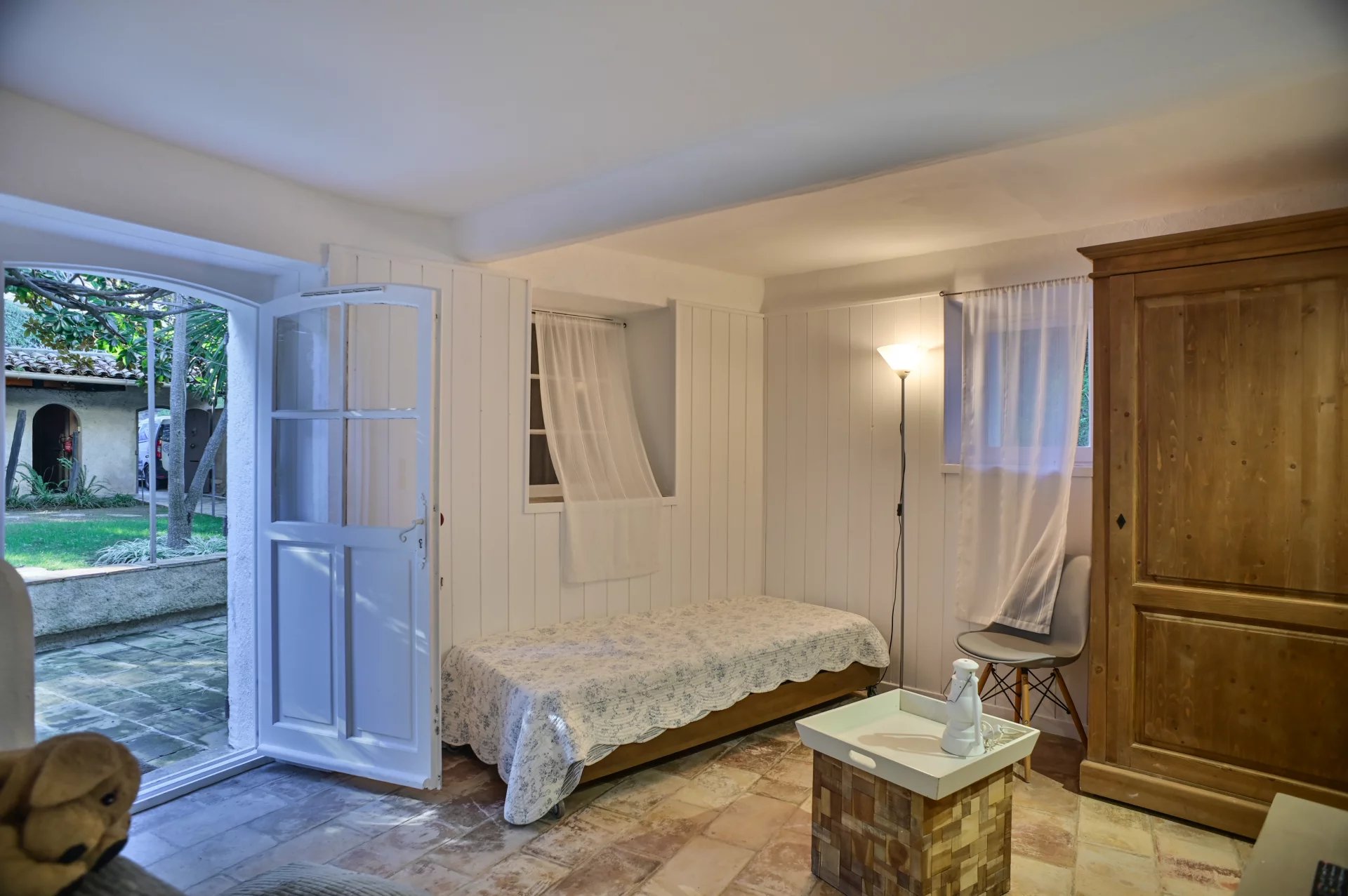 Intérieur d'une chambre avec un lit, une fenêtre ouverte sur le jardin et un meuble en bois.