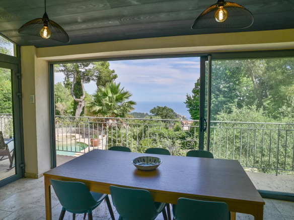Intérieur d'une villa à Èze avec vue sur la mer, table à manger et fenêtres ouvertes sur le jardin.