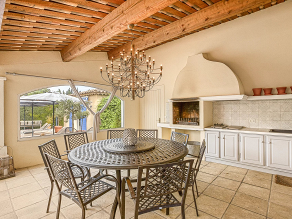Terrasse couverte avec table et chaises en métal, luminaires suspendus, barbecue intégré.