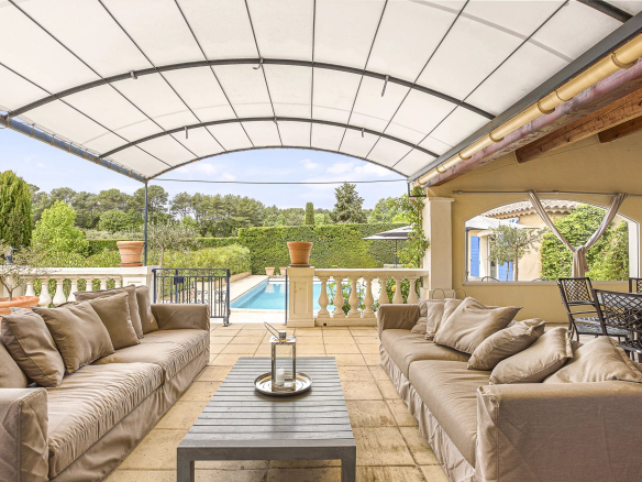 Terrasse couverte avec canapés et table, vue sur piscine et jardin paysager.